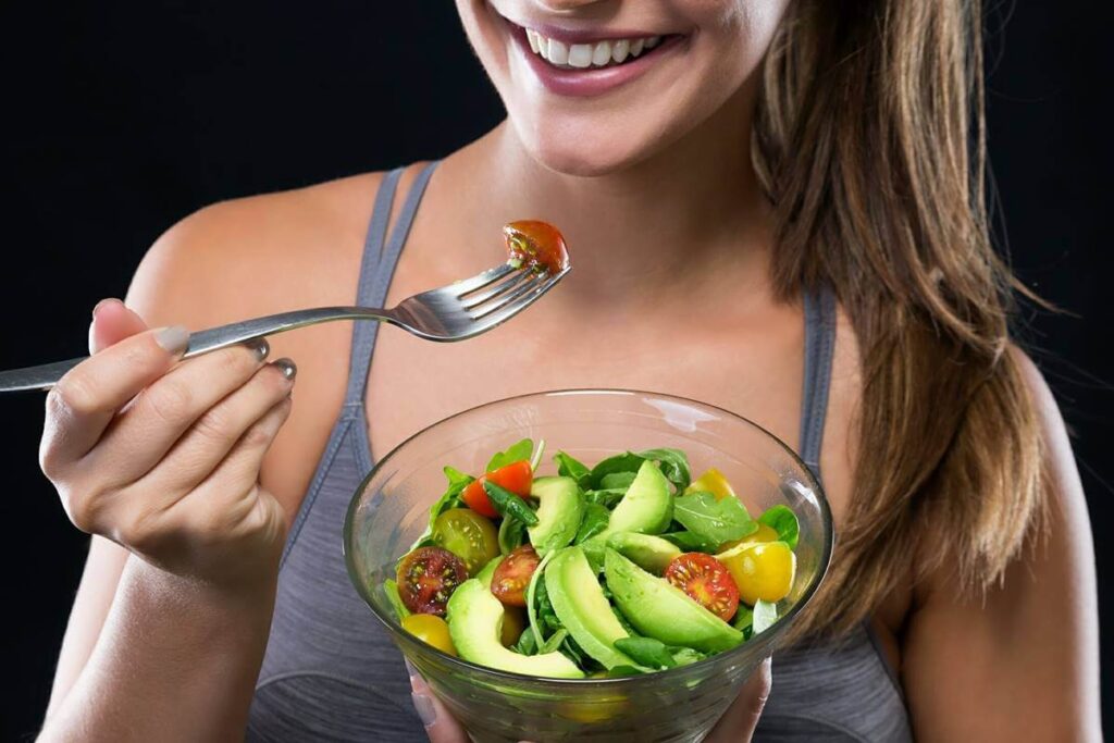 Visão geral: A imagem apresenta uma mulher com um grande sorriso, segurando uma tigela com salada e uma garfada com um tomate cereja. O fundo é preto, criando um contraste com o assunto, o que o destaca. O foco está no ato de comer a salada. A mulher: Rosto: A mulher está sorrindo, mostrando os dentes perfeitamente brancos. A boca está ligeiramente aberta e o sorriso parece genuíno e agradável. Os cantos de sua boca são mais claros do que o resto de sua face, possivelmente devido à iluminação. Cabelo: Seu cabelo é castanho e comprido, caindo sobre o ombro direito. Parece ter um estilo natural e está ligeiramente despenteado. Vestuário: Ela está vestindo uma regata cinza com alças finas. A roupa sugere um estilo de vida ativo. Tom de pele: A pele da mulher parece ligeiramente bronzeada. Mãos: Suas mãos são visíveis enquanto seguram a tigela e o garfo. Ela tem unhas curtas pintadas com esmalte cinza claro. A Salada: Tigela: A salada é servida em uma tigela de vidro transparente. Ingredientes: A salada consiste em folhas verdes escuras e vivas, possivelmente espinafre ou rúcula, fatias de abacate cremoso e tomates cereja de cor vermelha e amarela. Os ingredientes parecem frescos e coloridos, o que transmite uma sensação saudável. Montagem: A salada é empilhada na tigela. O abacate se destaca como o ingrediente mais proeminente, juntamente com os tomates cereja, que estão espalhados pela parte superior. Fatia de tomate: Um tomate cereja vermelho e brilhante é visto na ponta de um garfo de metal, pronto para ser comido. Foco: A combinação de seu sorriso radiante e da comida saudável cria uma atmosfera positiva e vibrante. A imagem busca comunicar uma sensação de bem-estar e alegria em relação à comida saudável. Iluminação e fundo: A imagem parece ter boa iluminação, fazendo com que as cores da salada e o sorriso da mulher se destaquem. O fundo preto destaca a mulher e a salada na imagem. O foco está nitidamente na mulher, na salada e no tomate na ponta do garfo. Composição: A mulher está centralizada e o visor é enquadrado na altura de sua face, com a tigela de salada e a garfada visíveis em primeiro plano. A combinação de cores e elementos na foto cria uma composição equilibrada e agradável, transmitindo uma sensação de saúde, bem-estar e alegria. Palavra chave: alimentação saudavel
