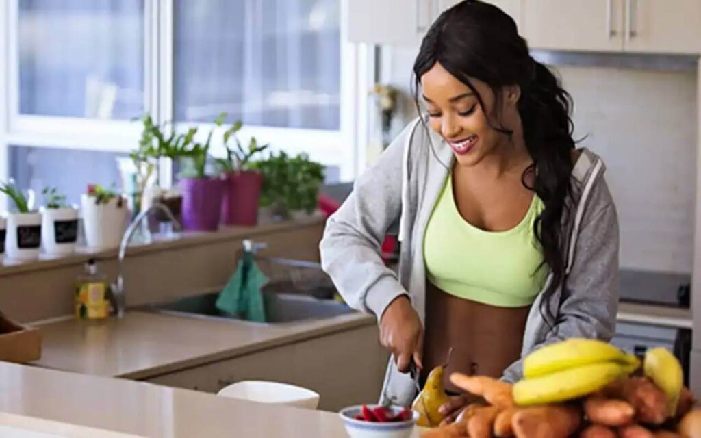 A imagem mostra uma mulher jovem na cozinha cortando frutas e legumes. Ela tem pele morena e está usando um top esportivo verde limão, uma jaqueta esportiva cinza e o cabelo escuro preso em um rabo de cavalo. Ela está sorrindo enquanto corta uma pera com uma faca. Em frente a ela está uma tigela com morangos cortados e várias frutas e legumes crus em um monte na bancada, incluindo bananas, batata doce e cenoura. A cozinha é brilhante e bem iluminada. Tem uma janela grande com várias plantas em vasos em uma borda. A pia está ao lado da mulher, com uma toalha verde pendurada no lado. As paredes e armários são de cor creme. Há um piso brilhante cor de creme que combina com as bancadas. A imagem sugere uma vida saudável e ativa, com foco em uma alimentação nutritiva. A mulher parece feliz e satisfeita enquanto prepara sua comida. A imagem tem um tom positivo, com cores brilhantes e uma composição nítida. Palavra chave: emagrecimento saudável