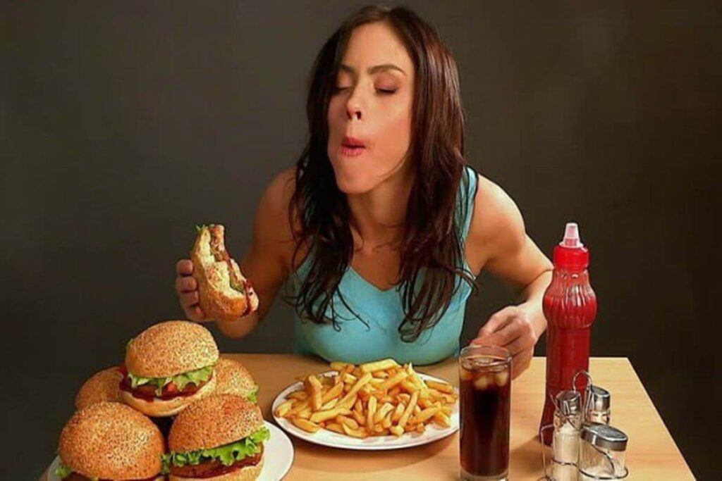 Visão geral A imagem mostra uma mulher sentada à mesa diante de uma abundância de junk food. Ela está no ato de comer um hambúrguer e parece bastante desfrutar de sua refeição. O cenário e a iluminação dão à imagem uma sensação um pouco estilizada e produzida. Detalhes da mulher Aparência: A mulher tem longos cabelos castanhos, sobrancelhas marcadas e lábios bem definidos. Seus olhos estão fechados no momento em que ela está mordendo o hambúrguer. Ela parece estar no final dos 20 ou início dos 30 anos. Vestuário: Ela está vestindo uma regata azul claro, que sugere uma sensação casual, mas ligeiramente atlética. Seus ombros e braços são visíveis, indicando que ela é atlética ou que tem um bom nível de condicionamento físico. Expressão: A expressão dela é de puro prazer, com a boca bem aberta e os olhos fechados. Junk food Hambúrgueres: Uma pilha de vários hambúrgueres com pães dourados com gergelim é exibida em uma placa branca, com seus recheios (provavelmente alface, tomate e hambúrguer) visíveis. Batatas fritas: Há um prato com uma grande porção de batatas fritas douradas. Bebida: Um copo cheio de refrigerante com gelo é colocado à direita. Condimentos: Um frasco de ketchup e um conjunto de saleiro e pimenteiro em aço inoxidável são visíveis na parte direita da mesa. Comida nas mãos: A mulher está segurando um hambúrguer que parece ter sido mordido, revelando seu interior. Mesa e fundo Mesa: A mesa é de madeira clara com um acabamento liso. Fundo: O fundo é um fundo cinza escuro, quase plano, que garante que o foco seja inteiramente sobre a mulher e a comida. Iluminação e cores Iluminação: A iluminação é uniforme, mas com uma leve sensação de luz de estúdio, criando um ambiente um pouco artificial. Cores: As cores são saturadas, com os tons dourados e castanhos da comida contrastando com a camisa azul claro e o fundo cinza-escuro. Impressão geral A imagem provavelmente visa transmitir uma sensação de prazer, indulgência e talvez até mesmo exagero no consumo de alimentos não saudáveis. A combinação de expressão facial da mulher, a grande quantidade de comida e o fundo estilizado dão à imagem uma sensação deliberadamente chamativa. A imagem poderia ser usada para temas como indulgência, indulgência na alimentação ou até mesmo como parte de uma crítica ao fast food. Palavra chave: inibidor de apetite